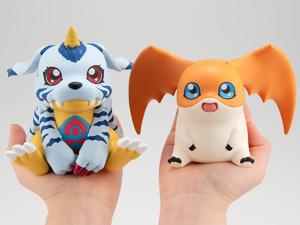 The same figure as previous, but being held up by hands. It is large enough to fully cover the persons palm. The other hand has a similar sized figure of Patamon, an orange and white hamster-shaped Digimon with large ear wings.